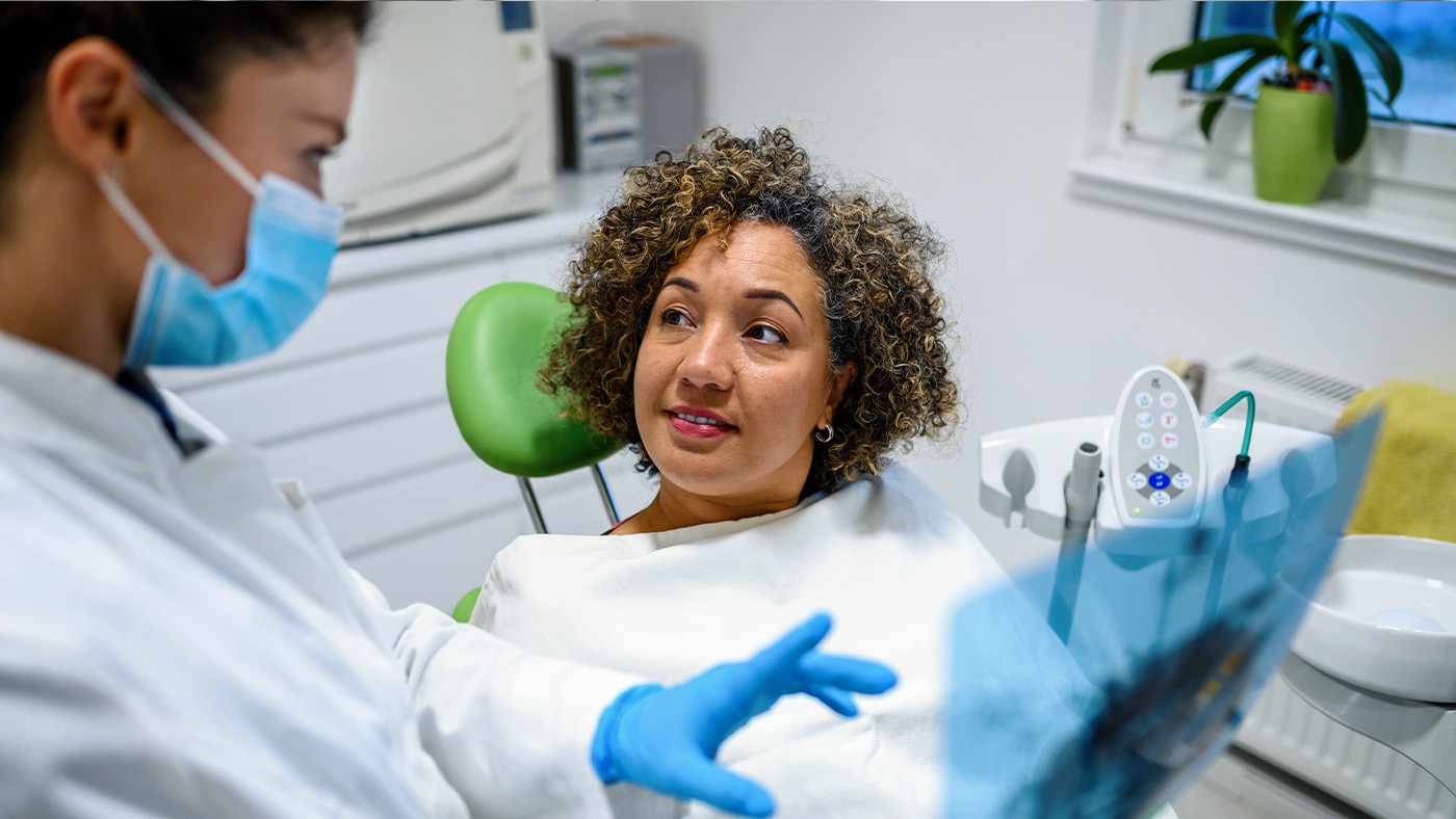 A woman sits in a dental chair while a masked dentist explains something, holding up an X-ray in a clinical setting.